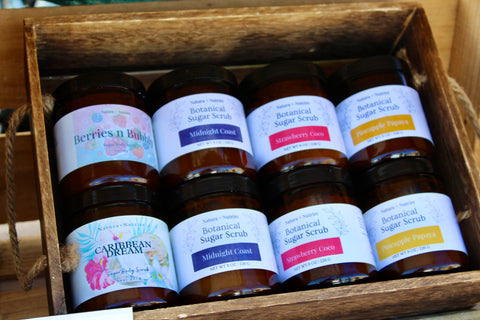 Eight Botanical Sugar Scrub jars in a wooden crate, showing Berries n Bubbly, Caribbean Dream, Midnight Coast, Strawberry Coco, and Pineapple Papaya scents