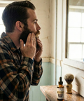 Man in a plaid shirt examining his beard in front of a mirror, with a bottle of Stoic Beard Oil and a brush on a wooden shelf