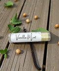 Vanilla Mint Olive Oil Lip Balm tube lying on a wooden deck surrounded by mint leaves, vanilla beans, and vanilla bean pods