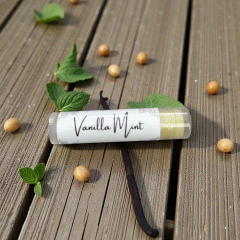 Vanilla Mint Olive Oil Lip Balm tube lying on a wooden deck surrounded by mint leaves, vanilla beans, and vanilla bean pods