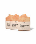 Three Strawberry Fields Handcrafted Soap bars wrapped in kraft paper sleeves, side by side on a white background