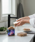 Hands lathering a bar of Luna Woods Handcrafted Soap under running water at a bathroom sink, with two more bars on the marble counter