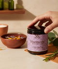 Hand opening a jar of Harmony Magnesium Cream on a beige tabletop with a lit candle, a bowl of dried lavender, and fresh rosemary sprigs