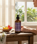 Harmony Magnesium Lotion pump bottle on a wooden table with a bowl of dried flowers, a folded embroidered cloth, and a woman seated by a sunlit window