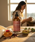 Enchanted Magnesium Lotion pump bottle on a wooden table with dried orange slices, hibiscus flowers, and eucalyptus, a woman standing near a sunlit window in the background