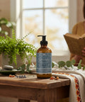 Serenity Magnesium Lotion pump bottle on a wooden table with a potted fern, eucalyptus sprigs, dried lavender, and a woman reading in the background