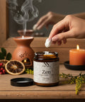 Hand dipping a cotton swab into an open jar of Zen Magnesium Cream on a wooden table with a lit candle, terracotta oil diffuser, dried orange slices, and red berries