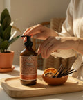Hands applying Bliss Magnesium Lotion at a wooden table with dried orange slices, vanilla beans, and a potted plant