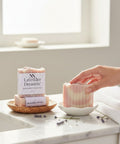 Hand reaching for a bar of Lavender Dreamin' Handcrafted Vegan Soap on a marble bathroom counter with fresh lavender sprigs and a white towel