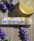 Lavender Lemonade Olive Oil Lip Balm tube with fresh lavender sprigs and a glass of lemonade on a woven placemat