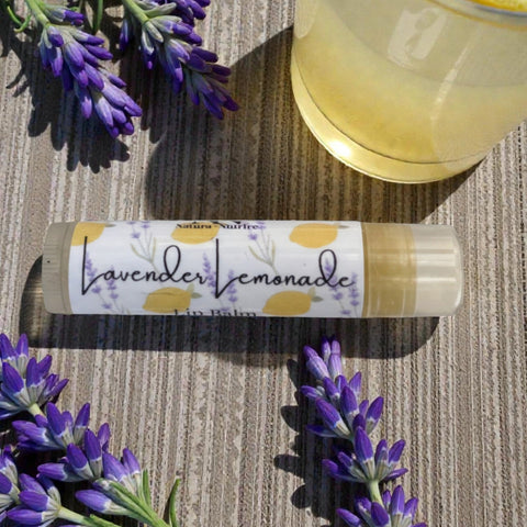 Lavender Lemonade Olive Oil Lip Balm tube with fresh lavender sprigs and a glass of lemonade on a woven placemat