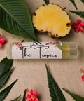 Tropics Pineapple Olive Oil Lip Balm tube surrounded by pineapple slices, tropical leaves, and hibiscus flowers on a beige background