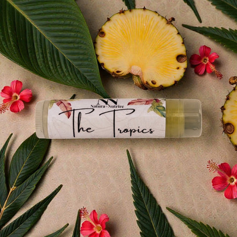 Tropics Pineapple Olive Oil Lip Balm tube surrounded by pineapple slices, tropical leaves, and hibiscus flowers on a beige background