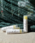 Three Vanilla Mint Olive Oil Lip Balm tubes standing and lying on a linen surface against a teal fabric backdrop
