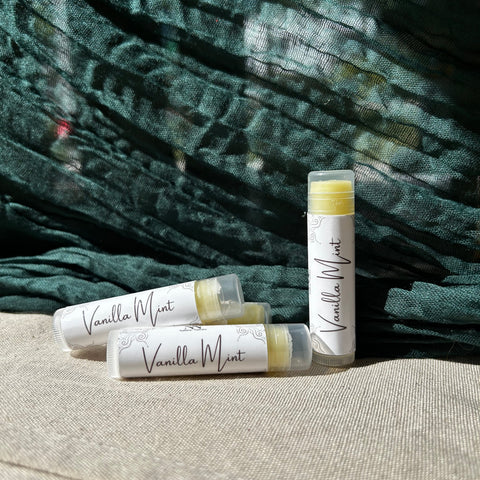 Three Vanilla Mint Olive Oil Lip Balm tubes standing and lying on a linen surface against a teal fabric backdrop