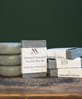 Charcoal & Tea Tree Face Bar Soap bars stacked on a wooden surface against a dark green background