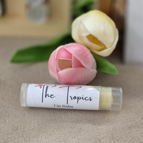 Tropics Pineapple Olive Oil Lip Balm tube labeled 'The Tropics' next to pink and yellow tulips on a linen surface