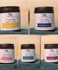 Foaming Sugar Scrub jars in five scents: Pineapple Papaya, Midnight Coast, Strawberry Coco, Watermelon Sugar, and Roxanne, on a teal fabric background
