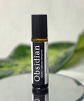 Obsidian Natural Botanical Fragrance rollerball bottle on a mirrored surface with green leaves in the background
