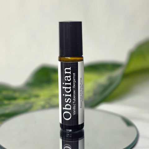 Obsidian Natural Botanical Fragrance rollerball bottle on a mirrored surface with green leaves in the background
