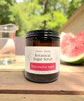 Botanical Sugar Scrub jar in Watermelon Sugar scent on a wooden table outdoors with fresh watermelon and a glass pitcher of water