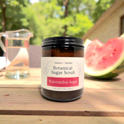 Botanical Sugar Scrub jar in Watermelon Sugar scent on a wooden table outdoors with fresh watermelon and a glass pitcher of water