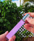 Hand holding the Lilac Handcrafted Non-Toxic Fragrance spray bottle against a lush green outdoor backdrop
