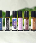 Natural Botanical Fragrances rollerball lineup: Seraph, Obsidian, Luna, Lilac, Roxanne, and Umber, on a white surface with green foliage in the background