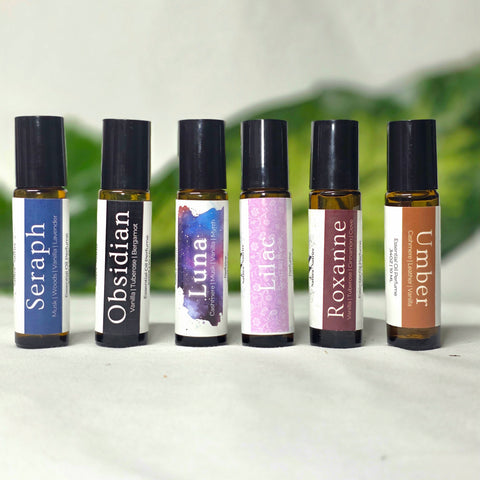 Natural Botanical Fragrances rollerball lineup: Seraph, Obsidian, Luna, Lilac, Roxanne, and Umber, on a white surface with green foliage in the background