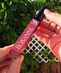 Hand holding the Roxanne Handcrafted Non-Toxic Fragrance rollerball bottle against a lush green outdoor backdrop