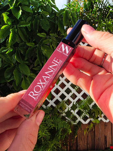 Hand holding the Roxanne Handcrafted Non-Toxic Fragrance rollerball bottle against a lush green outdoor backdrop