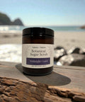 Botanical Sugar Scrub jar in Midnight Coast scent on a driftwood plank with a beach and rocky coastline in the background
