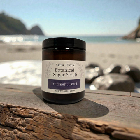 Botanical Sugar Scrub jar in Midnight Coast scent on a driftwood plank with a beach and rocky coastline in the background