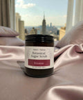 Botanical Sugar Scrub jar in Roxanne scent on pink satin bedding with a city skyline visible through the window