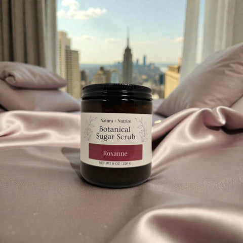 Botanical Sugar Scrub jar in Roxanne scent on pink satin bedding with a city skyline visible through the window