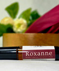 Roxanne Natural Botanical Fragrance rollerball bottle lying in front of a wooden bowl with white and pink flowers