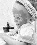 Black and white photo of a laughing toddler with Rose Renewal Clay Face Mask applied, sitting beside jars of skincare products