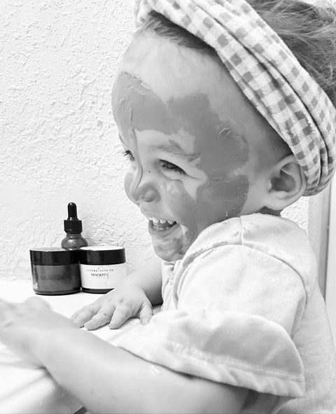 Black and white photo of a laughing toddler with Rose Renewal Clay Face Mask applied, sitting beside jars of skincare products
