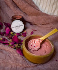 Rose Renewal Clay Face Mask jar with pink clay mask in a wooden bowl, scattered dried rose petals, and a wooden spoon on a mauve fabric background