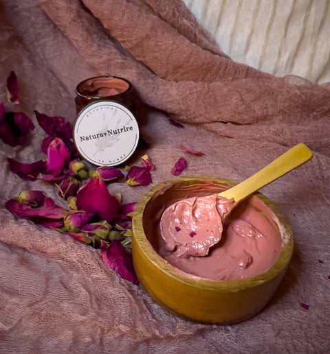 Rose Renewal Clay Face Mask jar with pink clay mask in a wooden bowl, scattered dried rose petals, and a wooden spoon on a mauve fabric background